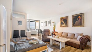 A modern living room with a sofa, a bed, a coffee table, and a chair. There are framed pictures on the wall and a plant on the coffee table.