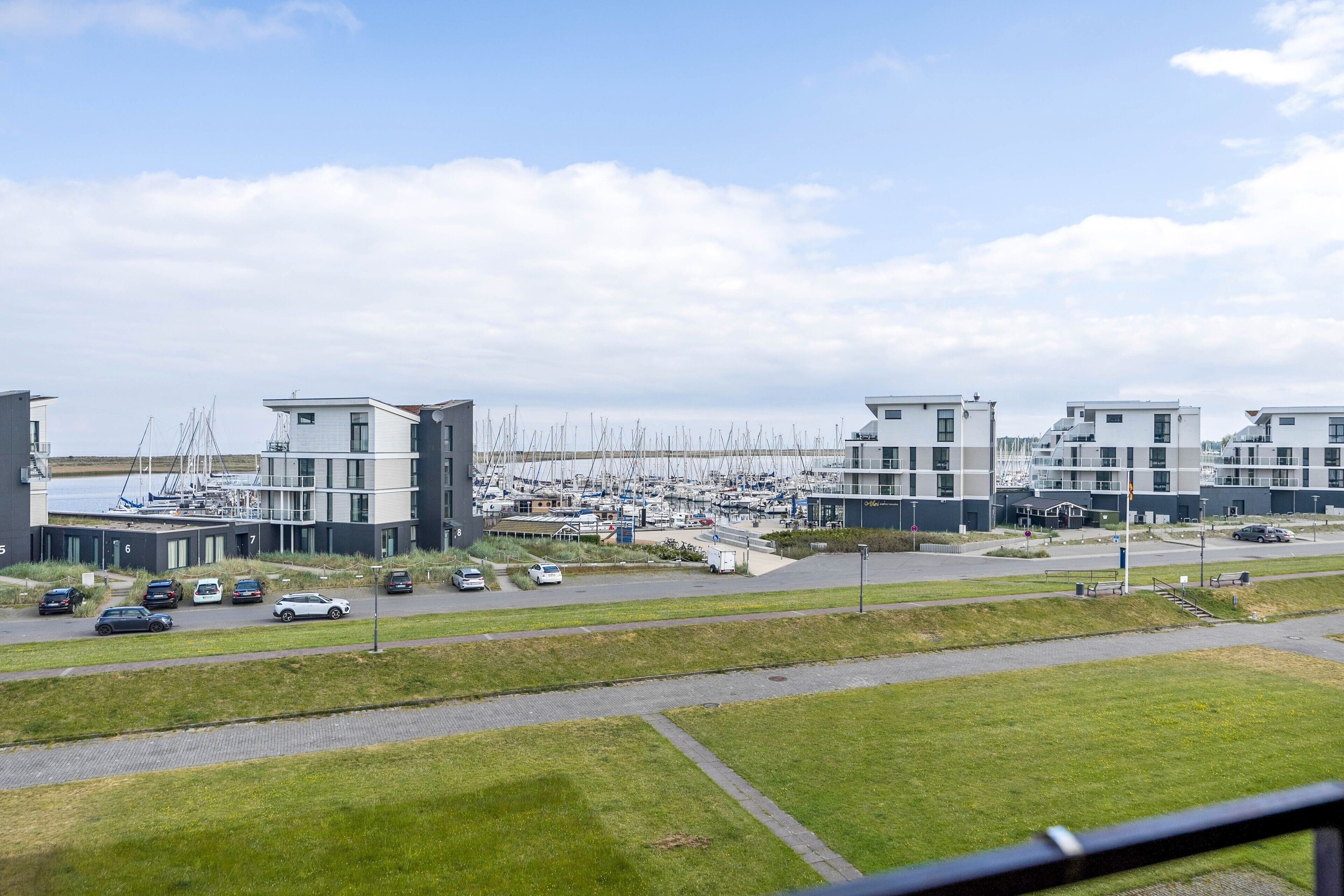 A marina with modern buildings, a parking area, and a grassy foreground.