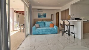 Un salon moderne comprenant un canapé, deux tabourets de bar, un coin cuisine et une porte-fenêtre coulissante donnant sur un balcon extérieur.