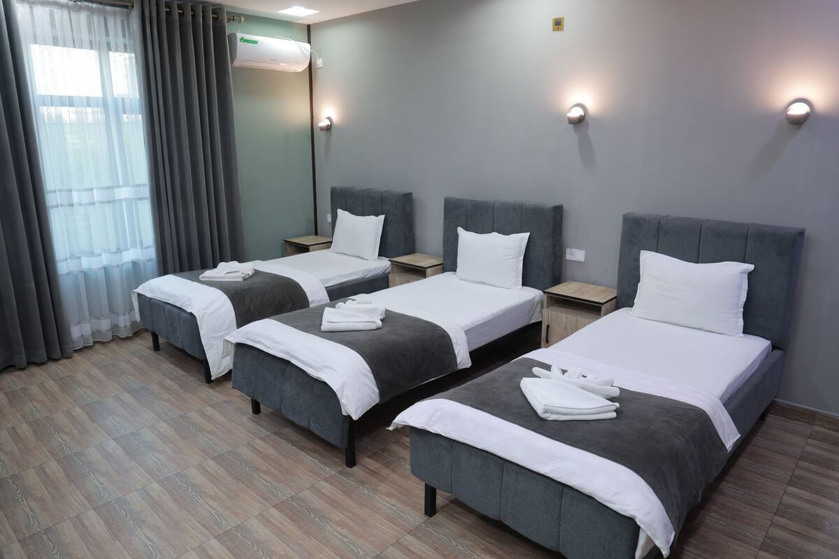 A hotel room with three beds, a wooden floor, a window with curtains, and a wall-mounted air conditioner.