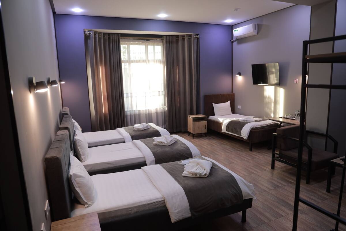 A hotel room with three beds, a desk, a chair, a TV, and a window with curtains.