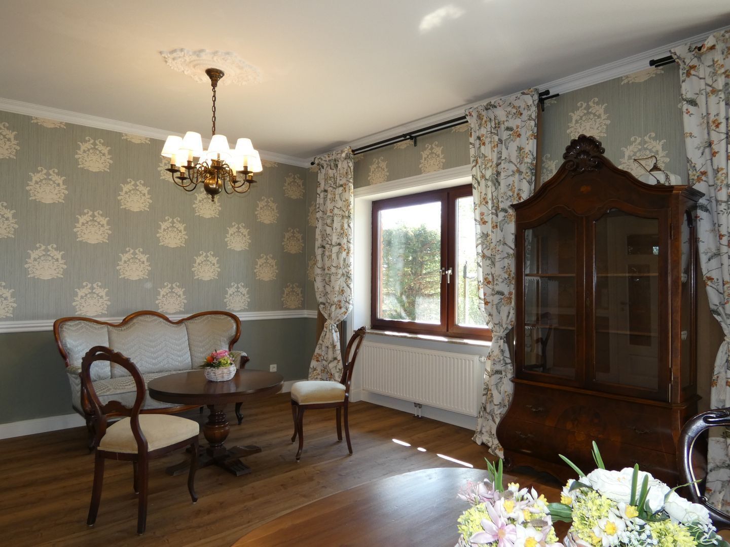 A room with a dining table, chairs, a chandelier, a cabinet, and a window with curtains.