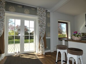 A room with a sliding glass door leading to a deck, a window with a basket of flowers, and two stools. - fewo1846 - Romance, Germany (Gelting)