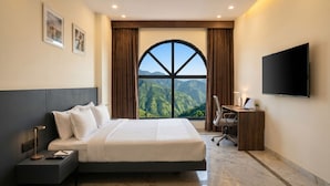 A hotel room with a large bed, a desk with a chair, a flat-screen TV, and a view of a mountainous landscape. - Eco Boutique Satva Udaipur - Vegetarian with all Options (Udaipur)