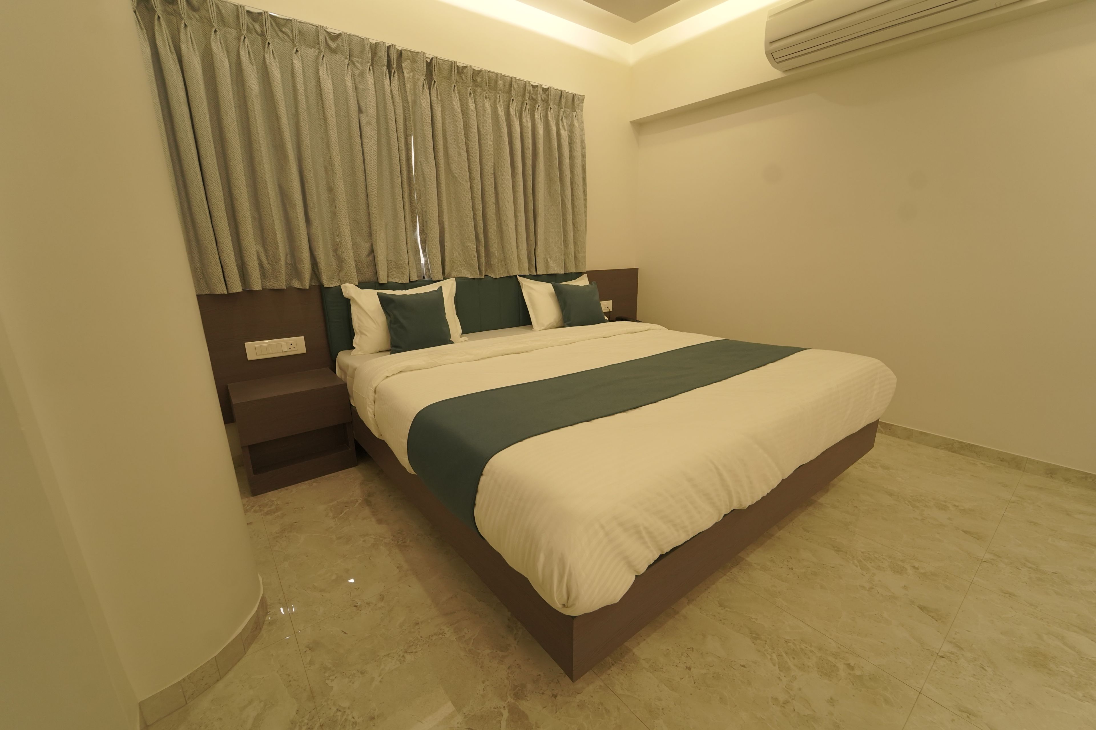 A hotel room with a bed, a nightstand, a power outlet, and a window with curtains.