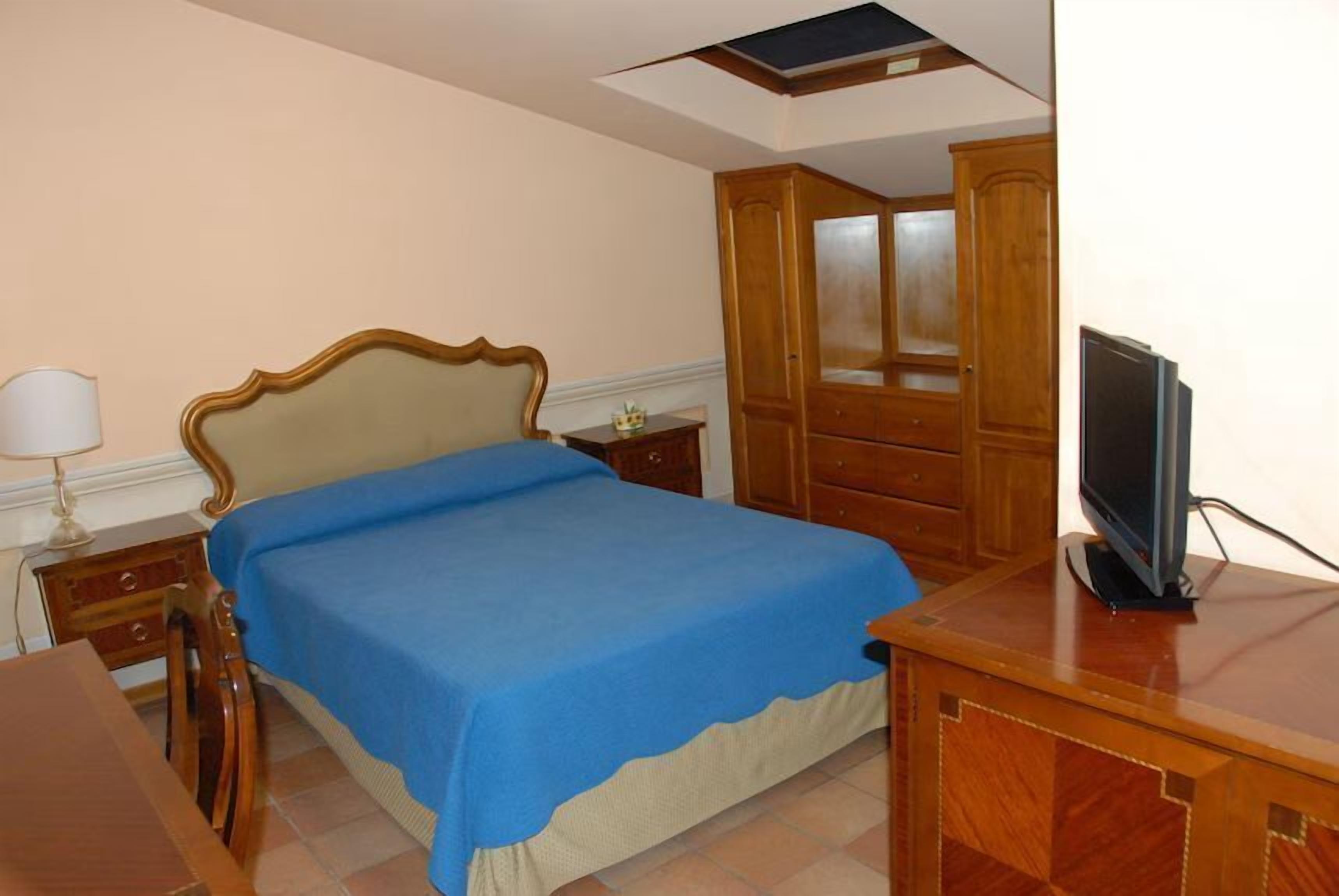 Un dormitorio con cama, armario de madera, escritorio con televisor y mesita de noche con lámpara.