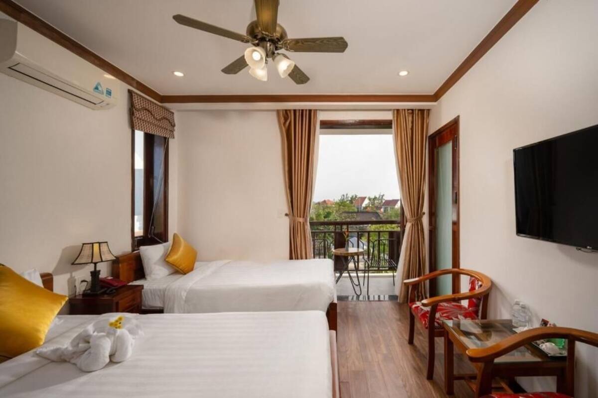 Hotelkamer met twee bedden, een plafondventilator, een televisie, een balkon met een tafel en stoelen, en een raam met gordijnen.