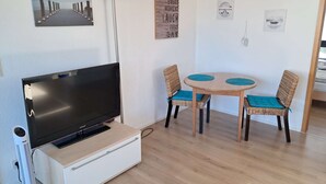 Una stanza con una TV a schermo piatto su un mobiletto bianco, un tavolo rotondo in legno con due sedie in vimini e una parete decorata con diverse immagini incorniciate.