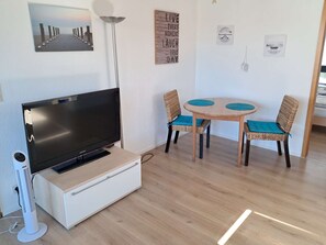 A room with a flat-screen TV on a white cabinet, a round wooden table with two wicker chairs, and a wall with various framed pictures. - Relax just 600m from the beach and only 100m from the forest (Kellenhusen)