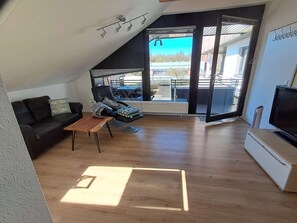 A modern living room with a black leather sofa, a wooden coffee table, a TV, and a large sliding glass door leading to an outdoor area. - Relax just 600m from the beach and only 100m from the forest (Kellenhusen)