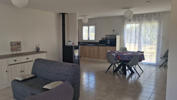 Una sala de estar con sofá, una mesa de comedor con sillas, una zona de cocina con estufa y un amplio ventanal con cortinas.