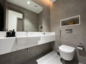 Ein modernes Badezimmer mit Waschbecken, Spiegel und Toilette, grauen Fliesen und einem Ablagefach für Pflegeprodukte.