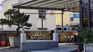 Un espacio al aire libre cubierto con mesas y sillas, un árbol en maceta y un edificio con depósito de agua y unidades de aire acondicionado.