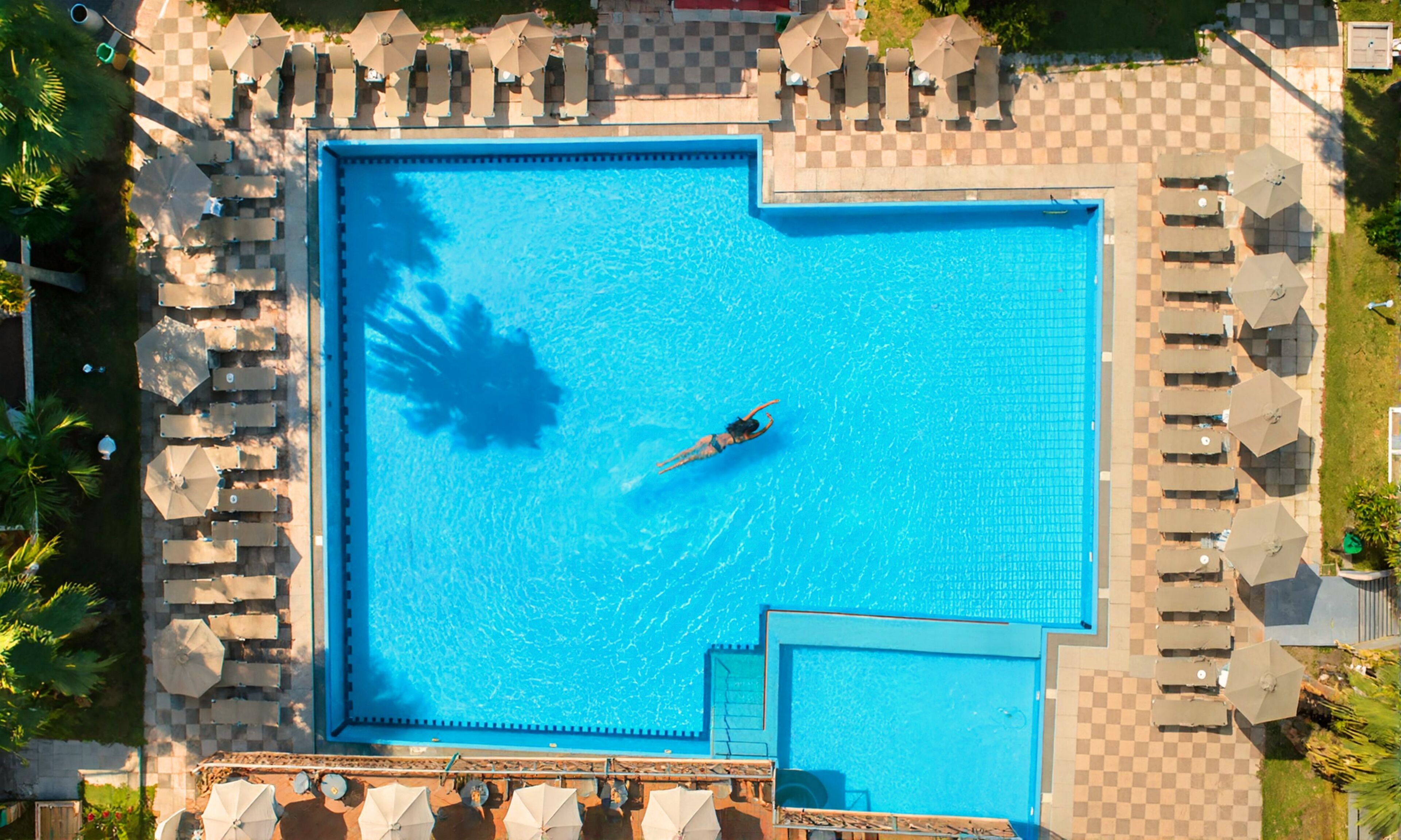 Ein großer, rechteckiger Swimmingpool mit einer darin schwimmenden Person, umgeben von Liegestühlen und Sonnenschirmen.