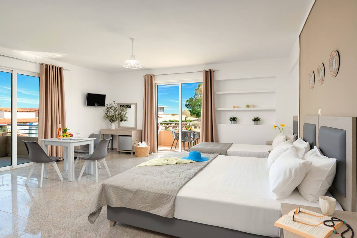 A hotel room with a balcony, a desk with a chair, two beds, a TV, and a small table with a vase and flowers.