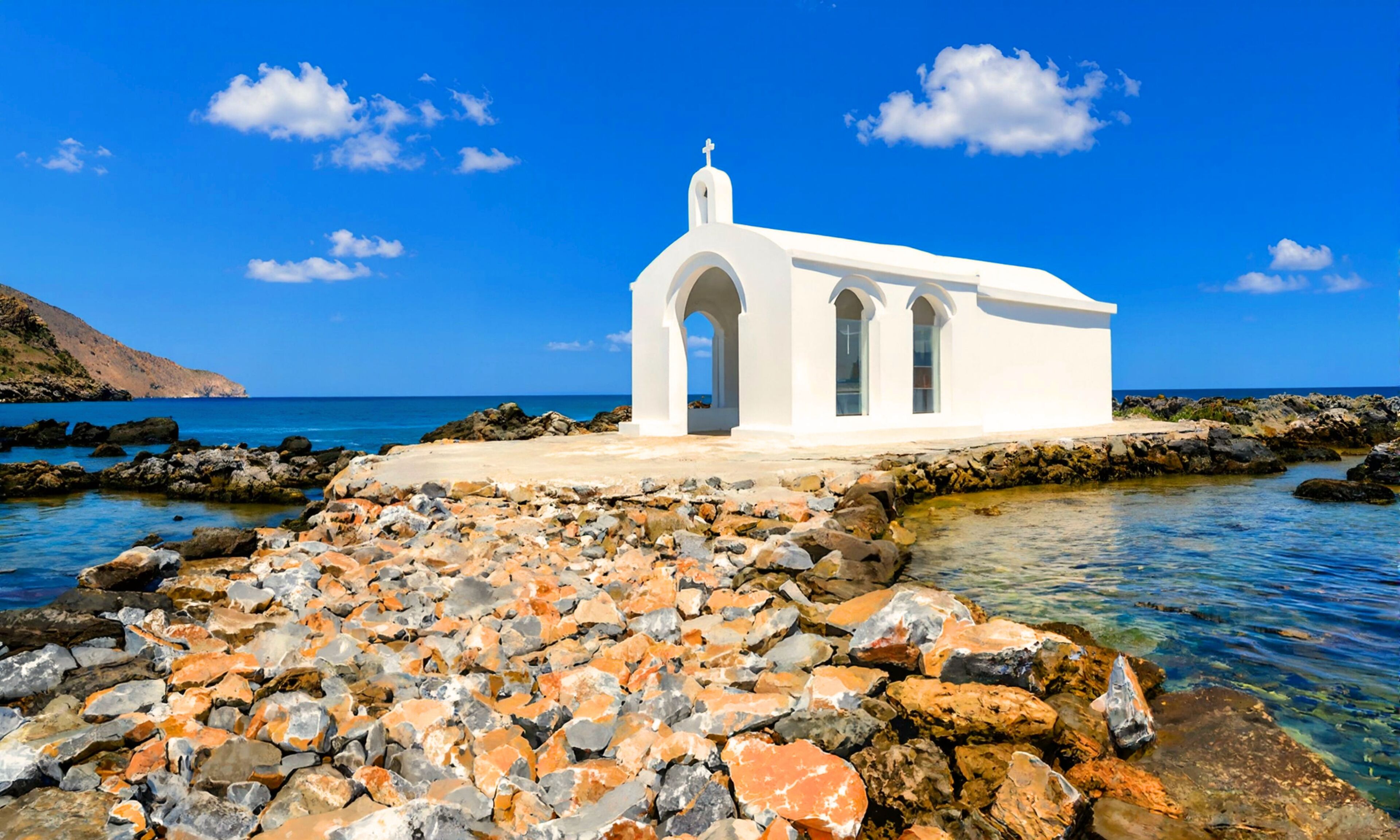 A white church with three arched windows and a cross on top, situated on a rocky shoreline with clear blue water and a distant mountain.