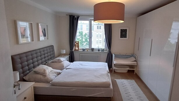 Een slaapkamer met een tweepersoonsbed, nachtkastjes, een lamp, een radiator, een raam met gordijnen en uitzicht op gebouwen.