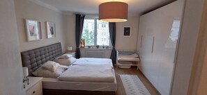 Camera da letto con un letto matrimoniale, comodini, una lampada, un termosifone, una finestra con tende e vista sugli edifici.