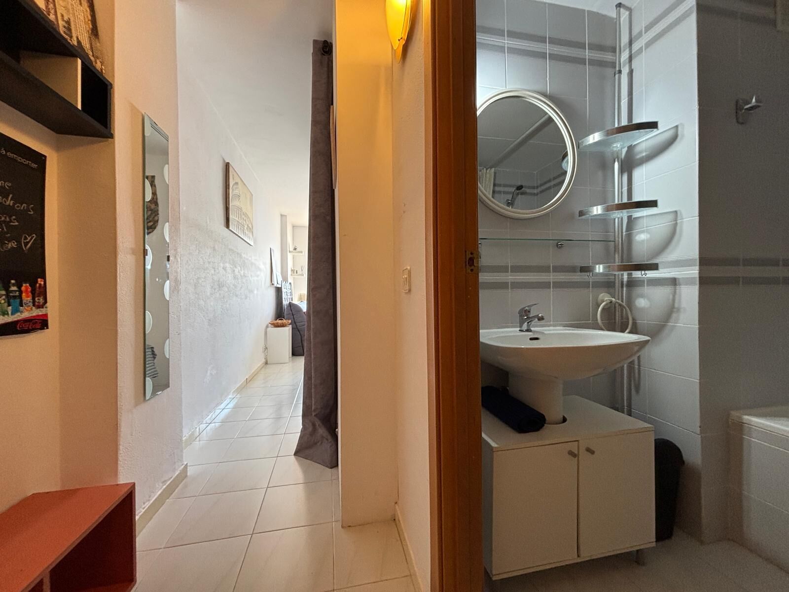 Une salle de bain avec un lavabo, un miroir et une douche. Un couloir mène à une autre pièce.