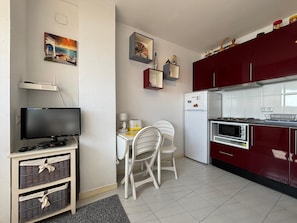 A compact kitchen with a refrigerator, microwave, and stove, a dining table with chairs, a television on a stand, and wall-mounted shelves with decorative items.