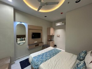 Et hotelværelse med seng, fjernsyn, spejl, ventilator, lille tekøkken og badeværelse.