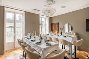 A dining area with a long table set for multiple guests, a chandelier, and large windows with a view of buildings outside. - Set of 4 apartments l 240m2 l Sleeps 14 l Ideal for receptions (Grenoble)