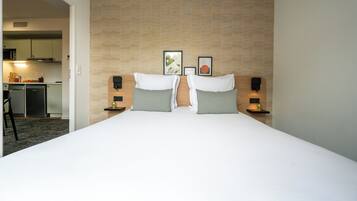 Una habitación de hotel con una cama grande, dos mesitas de noche, un cabecero con un papel tapiz estampado y una pequeña kitchenette al fondo.