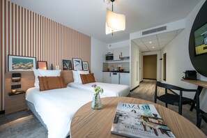 A hotel room with two beds, a dining table with a magazine, a kitchen area, and a hallway leading to another room. - Nemea Appart Hotel Nice Sunset Arenas (Nice)