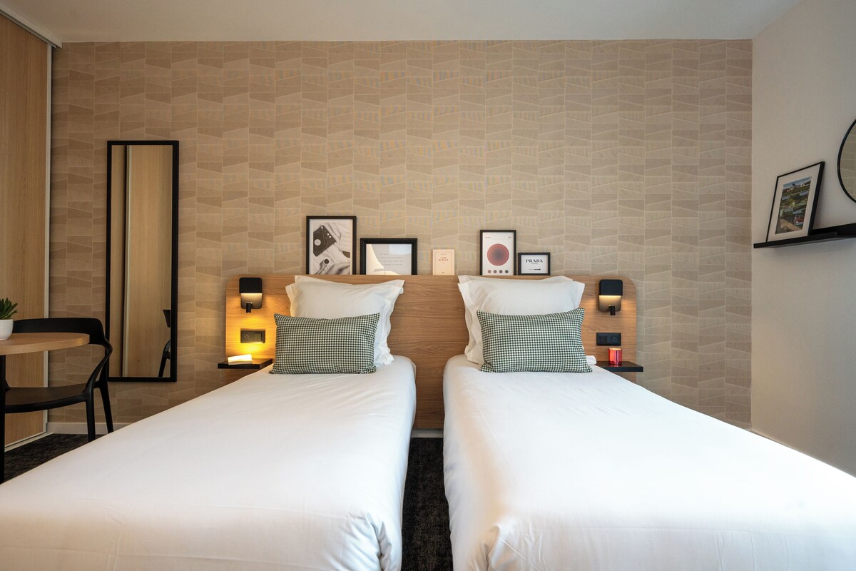 A hotel room with two beds, a small table, and a chair. There are framed pictures on the wall and a mirror.