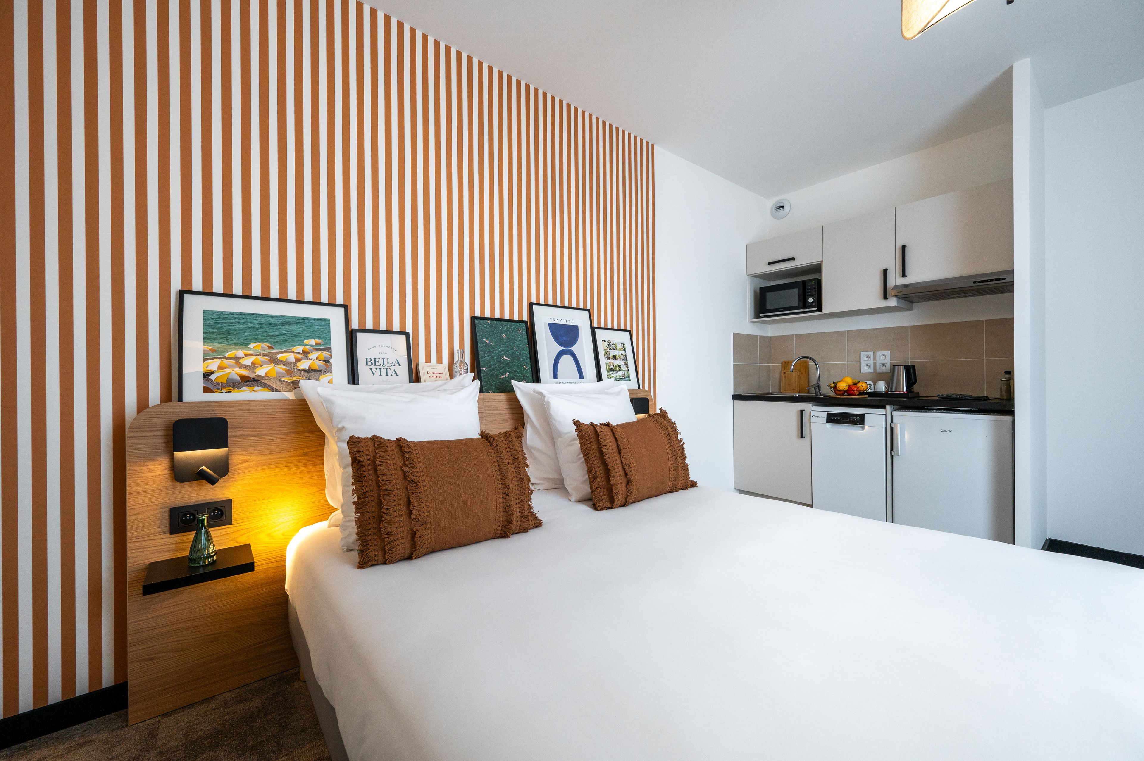 A hotel room with a bed, a bedside table with a lamp, a wall with striped wallpaper, and a kitchenette with a microwave and a refrigerator.