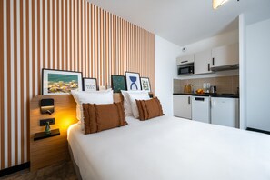 A hotel room with a bed, a bedside table with a lamp, a wall with striped wallpaper, and a kitchenette with a microwave and a refrigerator. - Nemea Appart Hotel Nice Sunset Arenas (Nice)
