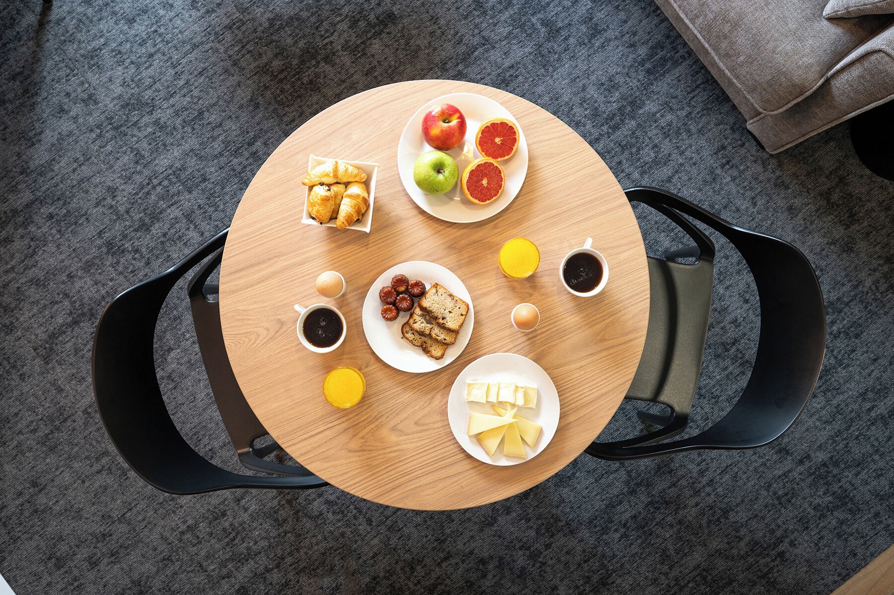 A round wooden table with breakfast items including croissants, fruits, and beverages, surrounded by black chairs.