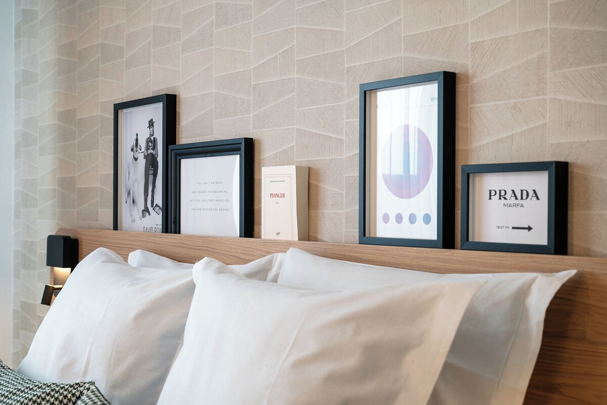 A hotel room with a bed, a wall with framed pictures, and a small table with a lamp.