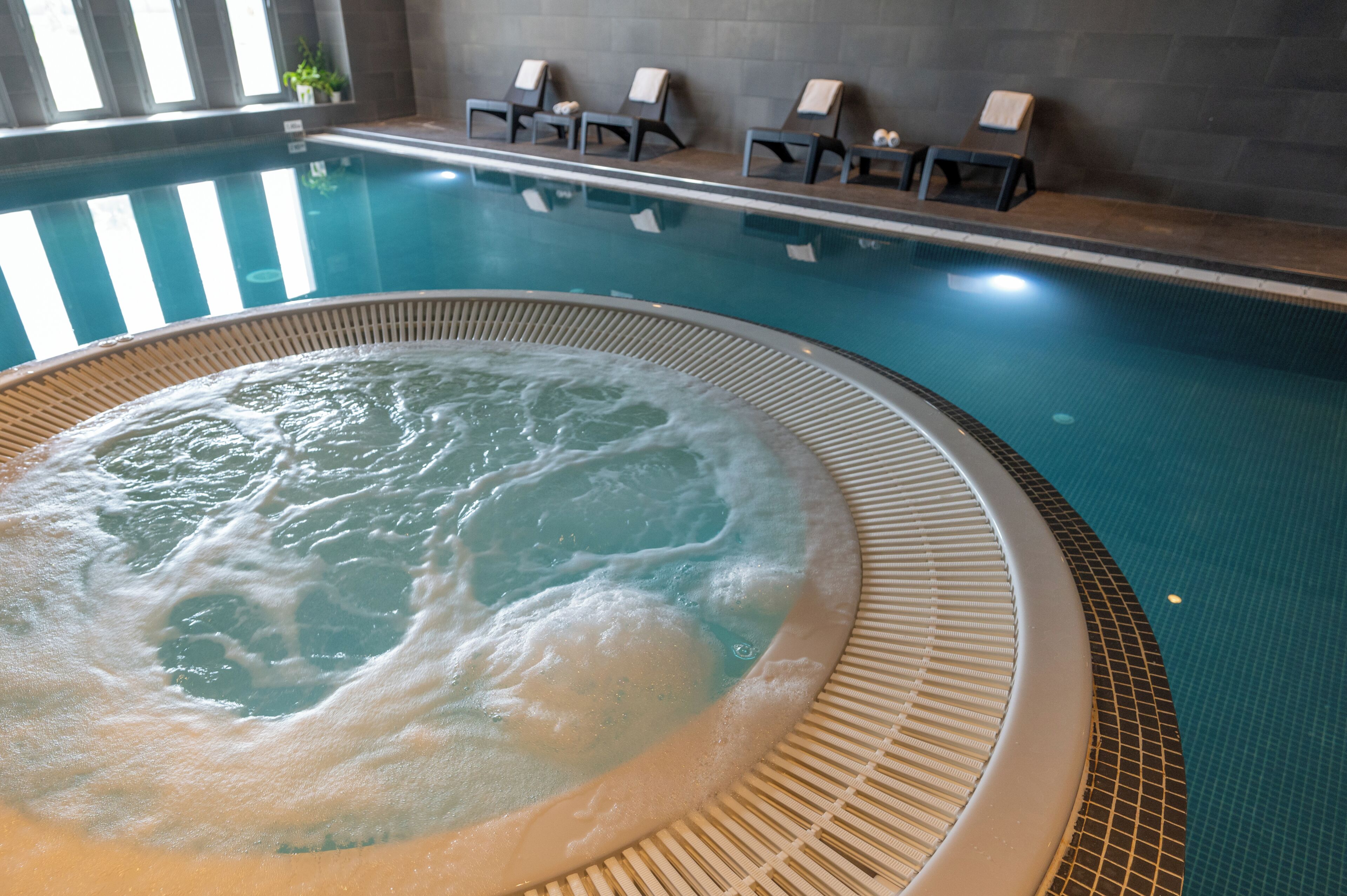 Un jacuzzi con borde de azulejos, una piscina con sillas de descanso y una pared revestida de azulejos.