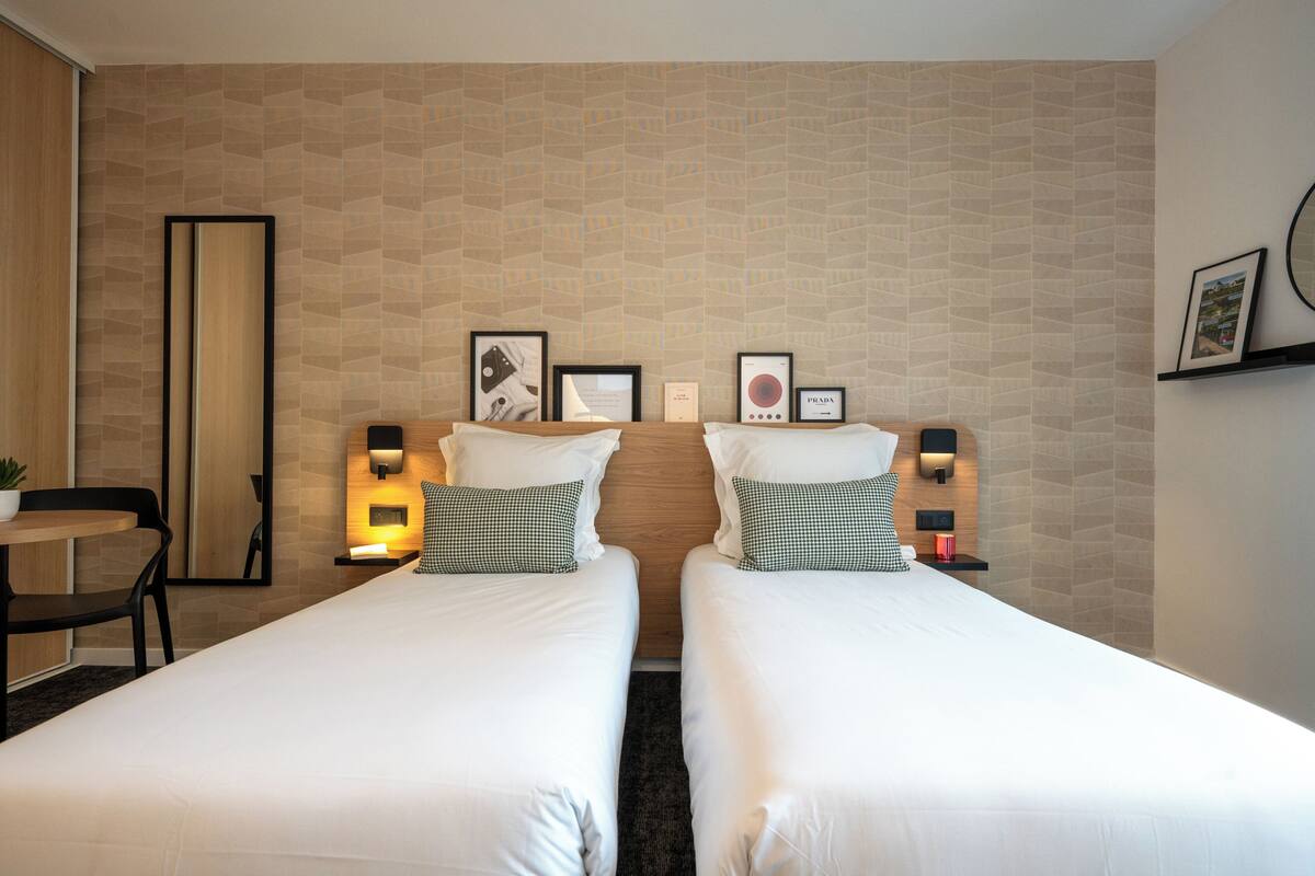 Quarto de hotel com duas camas, uma mesinha e uma cadeira. Há quadros nas paredes e um espelho.