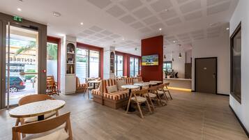 A hotel lobby with a reception desk, seating area with tables and chairs, and a large window overlooking the street.