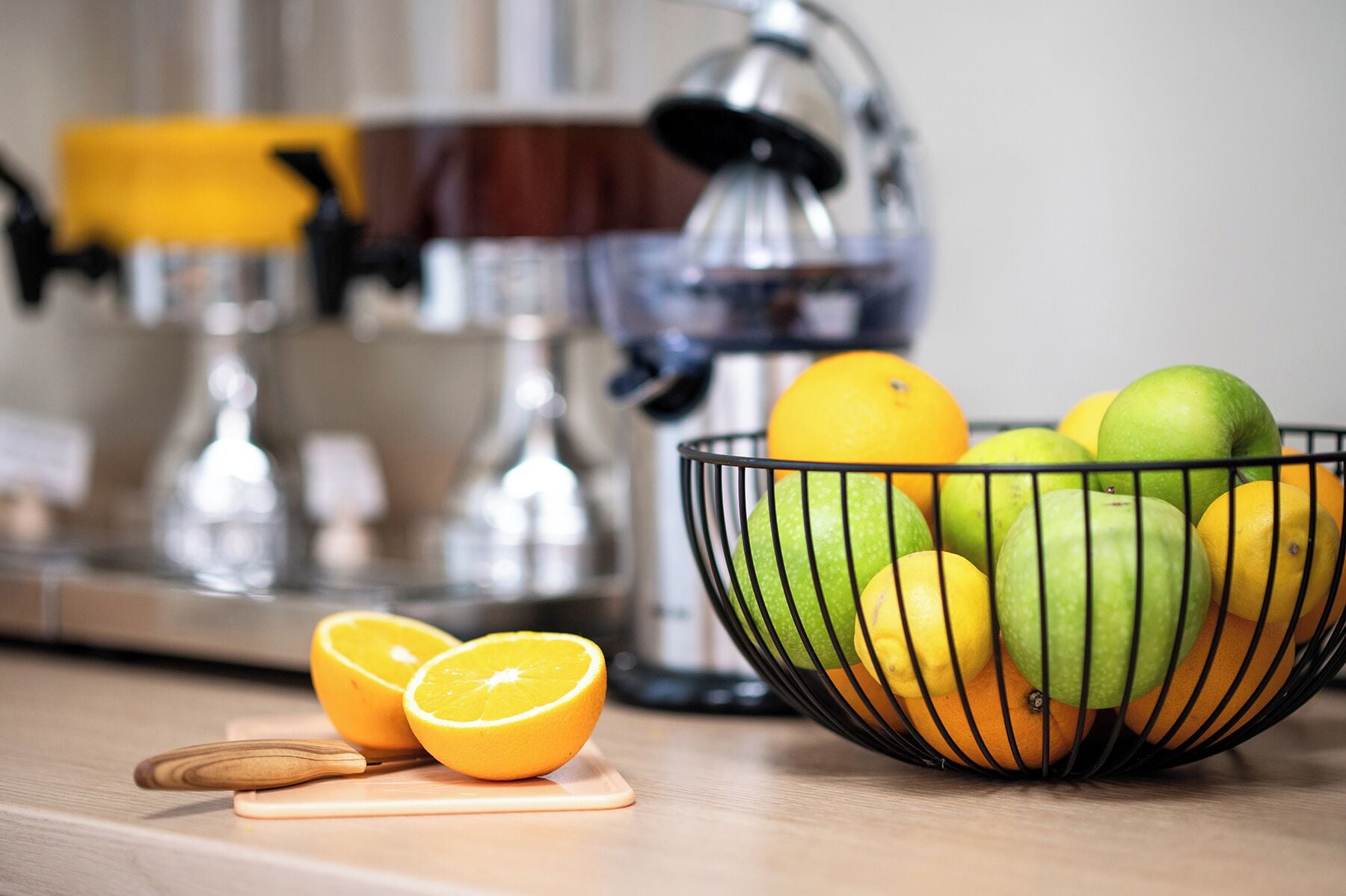 Eine Küchenarbeitsplatte mit einer Obstschale, die Orangen und Äpfel enthält, eine halbe Orange auf einem Schneidebrett und eine Kaffeemaschine im Hintergrund.