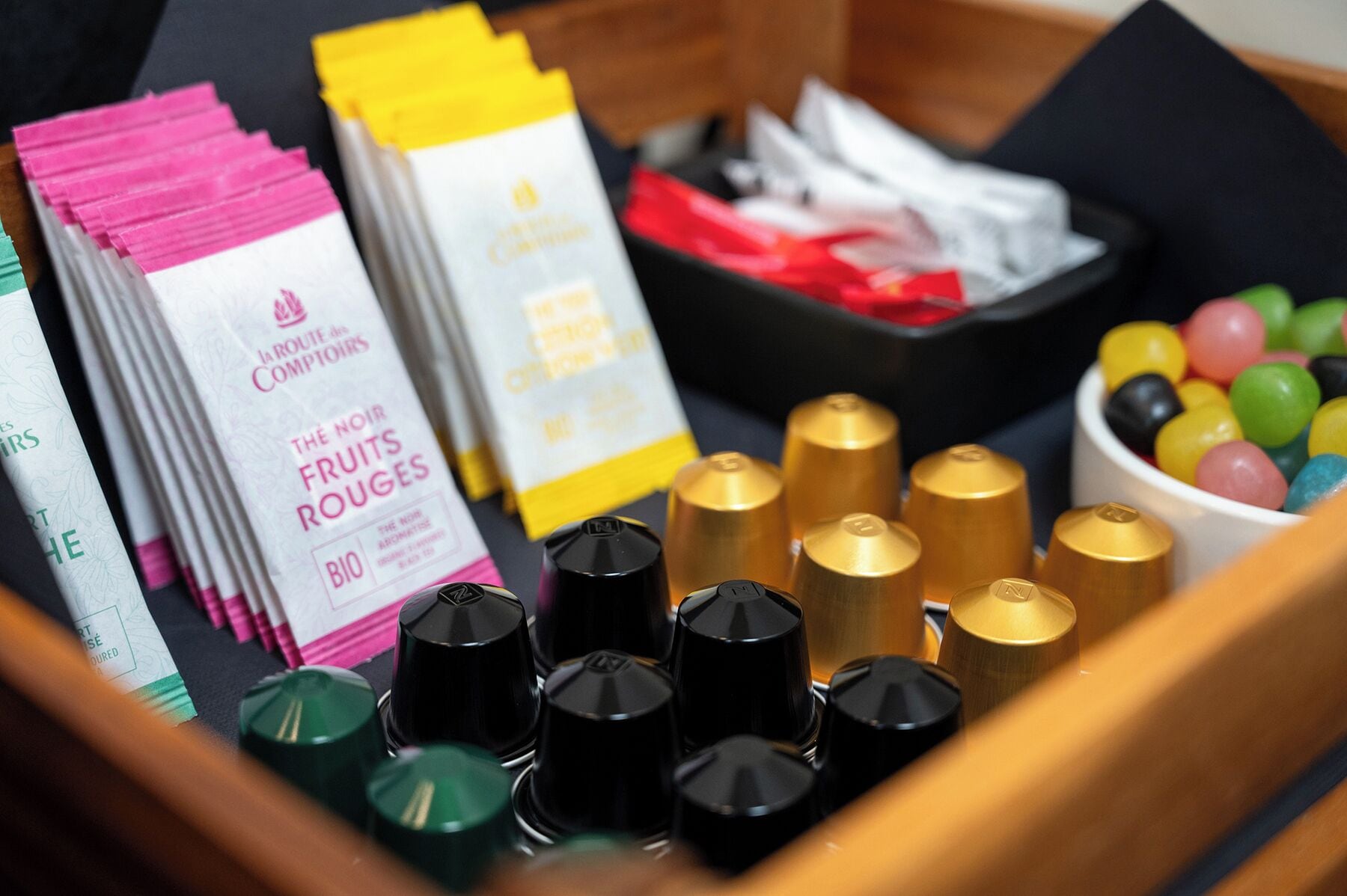 Una caja con varios artículos, incluyendo recipientes negros y dorados, cajas rosas y blancas con la etiqueta 'THE NOIR FRUITS ROUGES' y un tazón de bolas de colores.