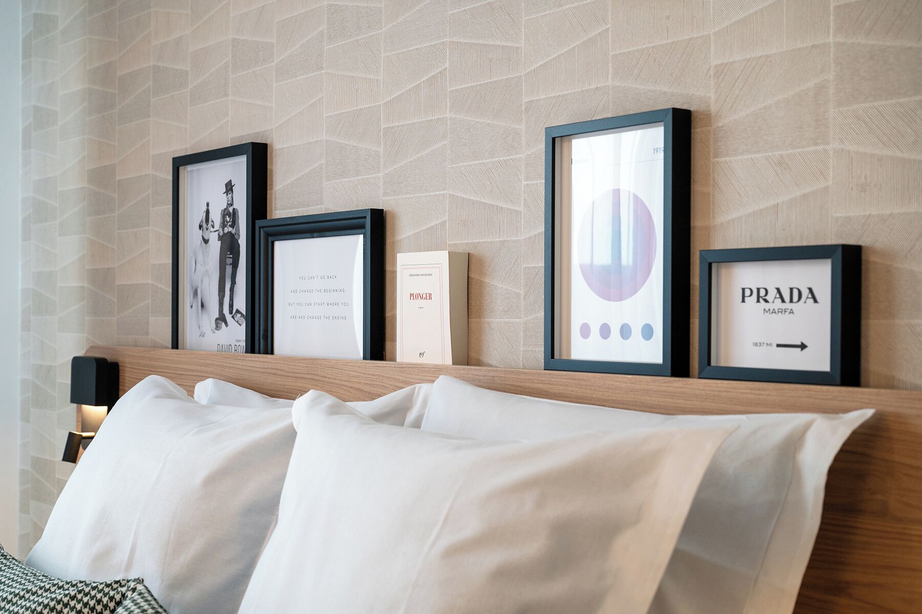 A hotel room with a bed, a wall with framed pictures, and a small table with a lamp.