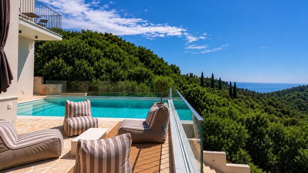 Zona junto a la piscina con sillones reclinables, barandilla de cristal y vistas a una ladera exuberante.