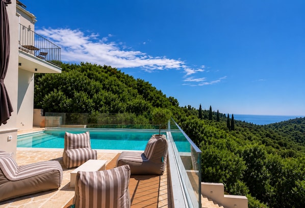 A poolside area with lounge chairs, a glass balustrade, and a view of a lush hillside. - Elegant Corfu Villa with Private Pool & Sea Views (Corfu)