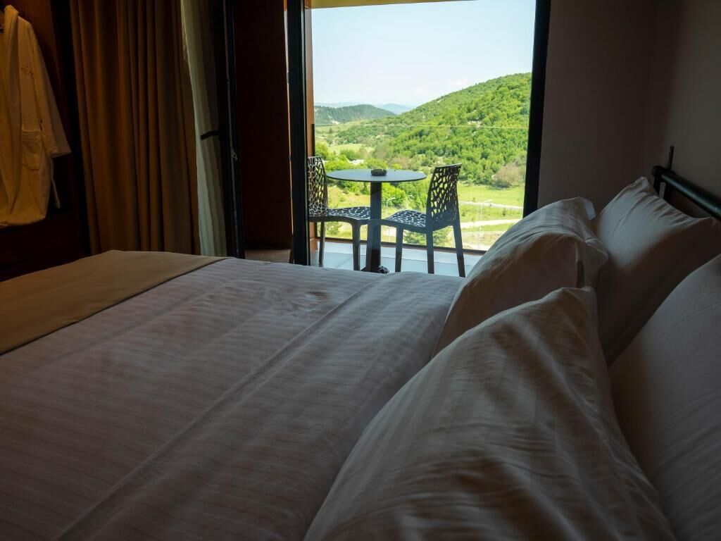 Quarto de hotel com uma cama, uma mesa com cadeiras e vista para uma paisagem verde pela janela.