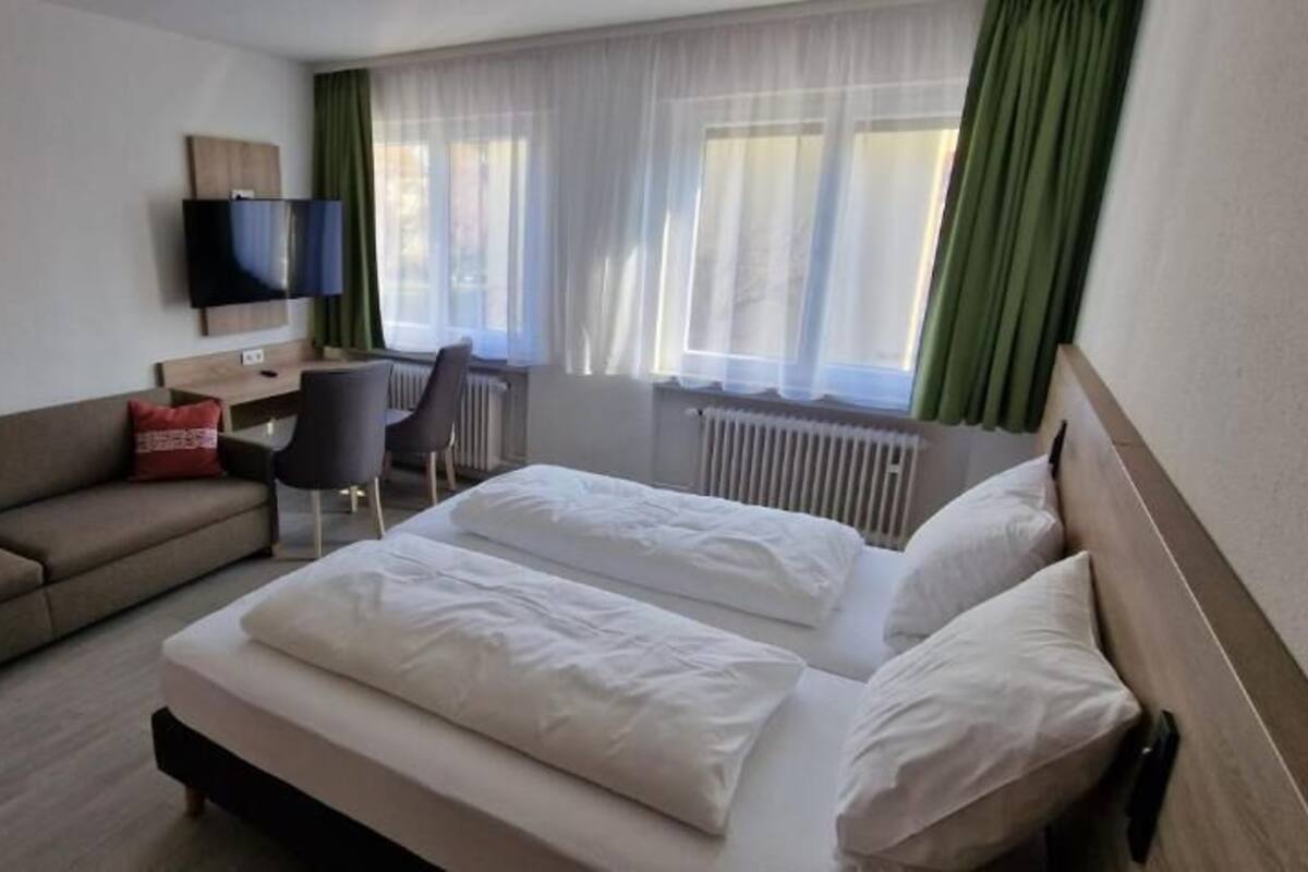 Habitación de hotel con cama, sofá, escritorio, silla, televisor y ventana con cortinas.