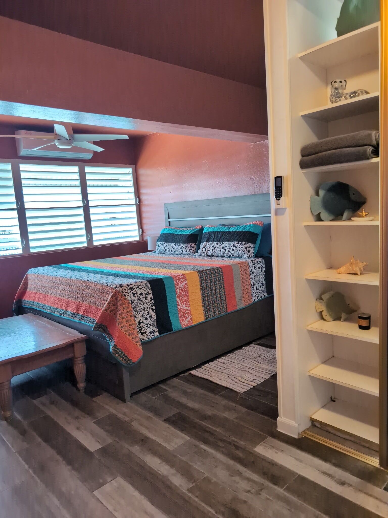A bedroom with a bed, a bench, a ceiling fan, a window with blinds, and a shelf with decorative items.