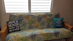 Un canapé orné de motifs de feuilles tropicales, deux coussins avec des inscriptions et une fenêtre avec des stores.