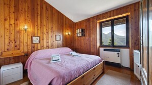 A wooden-paneled bedroom with a bed, bedside tables, a window with a mountain view, and a heater. - ‘Baita Piero e Afra’ Apartment with Mountain View, Balcony and Wi-Fi (Treviso Bresciano)