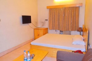 A hotel room with a bed, a small wooden table with bottles and cans, a TV mounted on the wall, and a window with curtains.