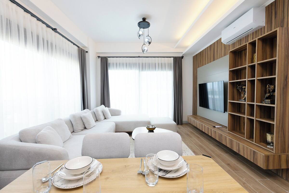 Un salon moderne avec un canapé d’angle, une table à manger dressée avec des assiettes et des verres, un téléviseur fixé au mur et une étagère en bois.
