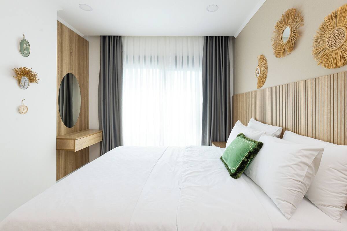 Une chambre d’hôtel avec un lit, une tête de lit en bois, un miroir et une fenêtre avec des rideaux.