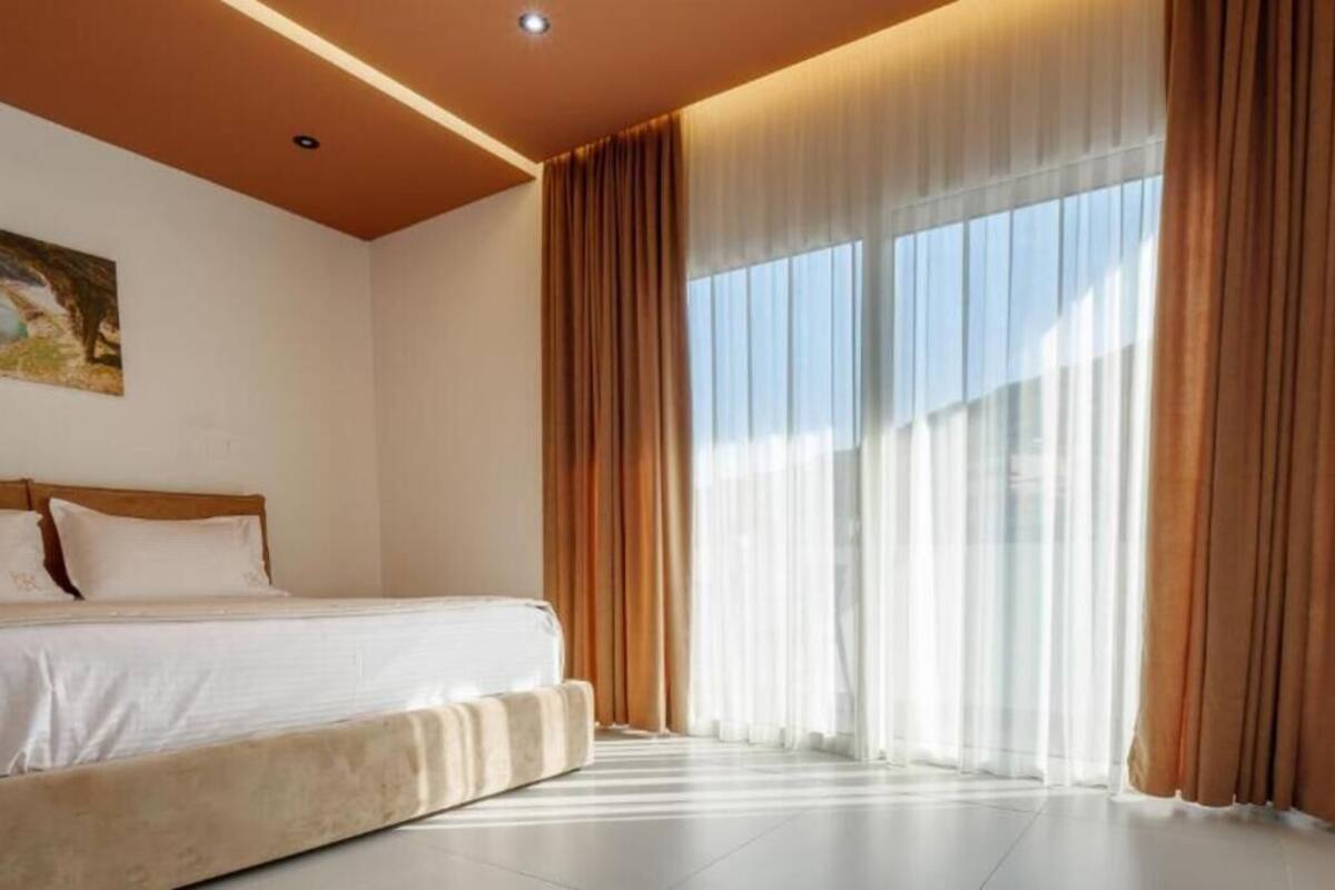 A hotel room with a bed, large windows with curtains, and a painting on the wall.
